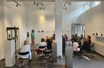 Groep mensen zit op klapstoelen in een witte galerie, luistert naar spreker tussen keramische kunstobjecten