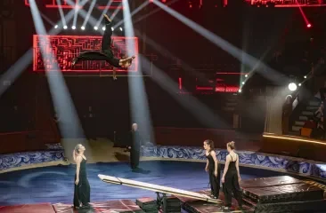 An acrobat leaps in the air above a balance beam.