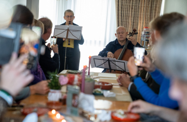 Musicians play the recorder and cello for a group of people seated at a long table in a living room.