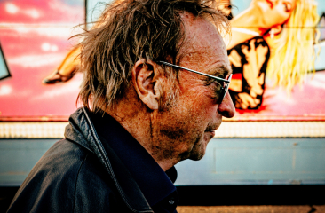 Een man met een onverzorgde haarsnit en zonnebril loopt langs een kleurrijke muur met een afbeelding van een vrouw.
