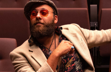 Een man met een baard en hoed zit ontspannen in een theaterstoel.