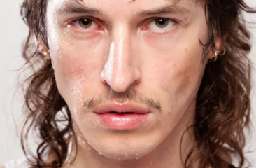 Close-up van een man met nat gezicht, lange donkere krullen en een dun snorretje, die intens in de camera kijkt.