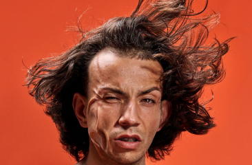 Portret van een man met wapperend haar en verwrongen gezichtsuitdrukking, tegen een feloranje achtergrond.