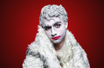 Persoon met wit geschminkt gezicht en haar, rode lippenstift en witte bontjas, poseert voor een rode achtergrond.