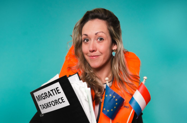Close up van Djûke Stammeshaus poserend voor een blauwe achtergrond. Ze draagt een oranje reddingsvest, een Europese en Nederlandse vlag en een uitpuilende map met migratie taskforce erop. 