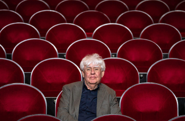Oudere man met grijs haar zit alleen op een rij in een lege zaal met rode theaterstoelen