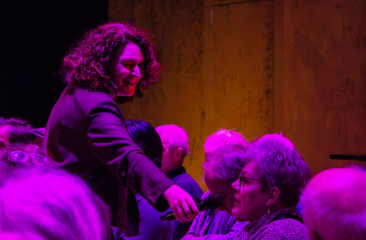 Vrouw met microfoon spreekt met iemand uit het publiek in een zaal met paarse en oranje verlichting