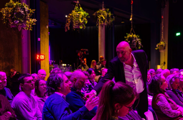 Een presentator in pak geeft een man uit het publiek een microfoon aan. Het publiek zit op stoelen in een roze verlichte zaal 
