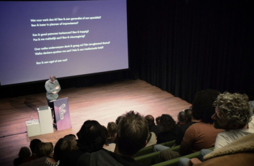 Spreker presenteert voor volle zaal in theater met projectiescherm en donkere gordijnen op de achtergrond