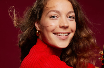 Vrouw met lang bruin haar en rode trui lacht naar de camera, houdt een klarinet vast voor een rode achtergrond