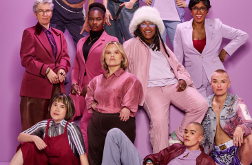 Groep van acht diverse mensen in kleurrijke outfits poseert voor een paarse achtergrond.