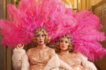 Promotiebeeld voor cabaretvoorstelling Harrekidee Plien en Bianca zitten naast elkaar en kijken bedenkelijk. Ze zijn gekleed in showgirl outfits: gouden glitterjurken, boa's en roze verentooien op hun hoofd. 