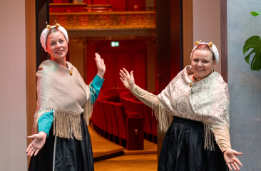 Twee vrouwen in traditionele klederdracht verwelkomen bezoekers bij een theateringang.  