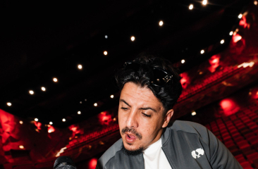 Foto van ICE op het podium. Hij draagt een zwart jack en heeft een microfoon in zijn hand. Achter hem zien we een lege theaterzaal met rode stoelen. 