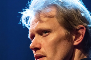 Close-up van een man met blond haar en serieuze blik, dramatisch belicht tegen een donkere achtergrond met blauw licht.