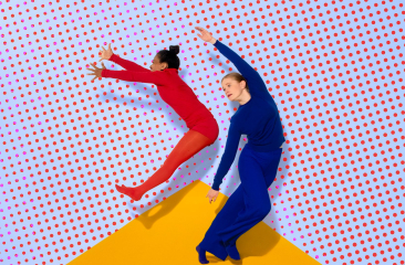 Twee dansers in rode en blauwe outfit springen voor een wand met rode stippen en een gele driehoek op de vloer