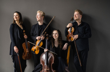Vier musici in zwarte kleding met strijkinstrumenten poseren ontspannen tegen een grijze muur