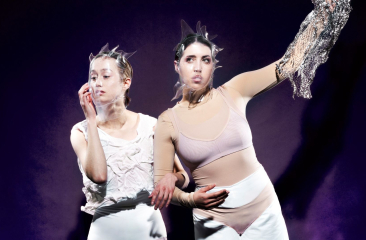 Twee performers in abstracte witte outfits en hoofdstukken poseren expressief tegen een paarse achtergrond