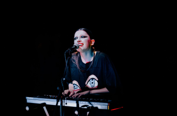  Artiest met opvallende make-up speelt keyboard en zingt op een donker podium, verlicht door een spotlight.