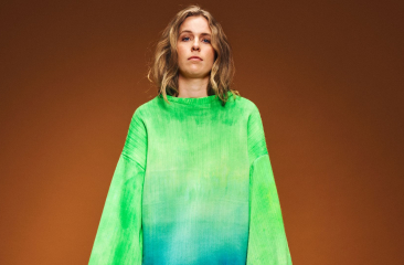 Model in oversized felgroene trui met blauw kleurverloop, poseert voor een effen bruine achtergrond.