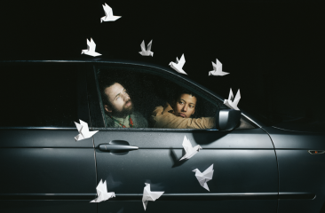 Twee personen zitten in een stilstaande auto terwijl witte papieren duiven rondom hen lijken te zweven in het donker