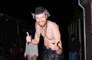 Lachende man met hoed en ontbloot bovenlichaam poseert met duimen omhoog tijdens een avondfeest.
