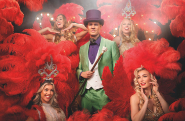 Tim Fransen is omringd door showgirls in outfits met rode veren. Tim draagt een groen pak met paarse hoed. 