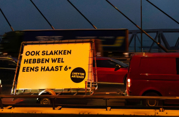 Verlichte aanhanger met tekst “Ook slakken hebben wel eens haast 6+” tussen stilstaand verkeer op brug in de avondspits