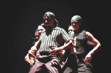 Groep dansers op een donker podium, intens belicht, met bandana’s en streetwear in een dynamische pose