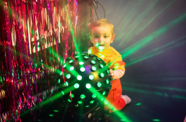 Peuter in oranje outfit kruipt bij discobal met groene lichtstralen en roze slierten op de achtergrond
