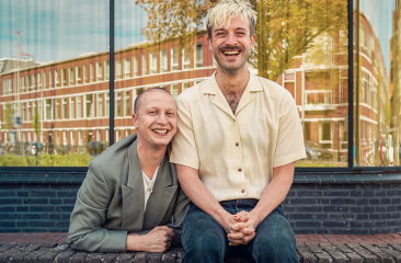 Twee lachende mannen zitten op een bakstenen muurtje voor een spiegelend raam met een stadspand op de achtergrond. De man links draagt een grijs pak, de man rechts een crèmekleurig overhemd en heeft blond haar.