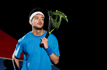 Man in blauw sportshirt met hoofdband en squashracket op een sportveld, zwarte achtergrond.