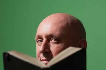 Kale man kijkt over een geopend boek richting camera, met groene achtergrond