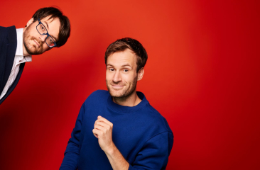 Promotiebeeld voor de cabaretshow Verbroedering. Roel en Jos Maalderink poseren voor een rode achtergrond. Jos draagt een wit overhemd en een donkerblauw colbert. Hij komt schuin het frame binnenkijken. Roel draagt een blauwe trui en lacht naar de camera. 