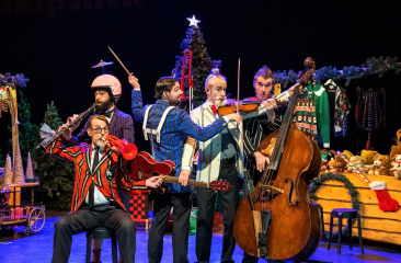 Vijf muzikanten in kleurrijke kleding bespelen verschillende instrumenten op een feestelijk versierd podium met een kerstboom en cadeaus op de achtergrond.