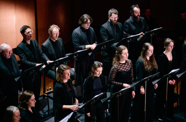 Een koor van mannen en vrouwen in zwarte kleding zingt vanaf muziekstandaards op een donker verlicht podium. Ze staan in twee rijen en concentreren zich op hun bladmuziek, met een houten wand op de achtergrond.