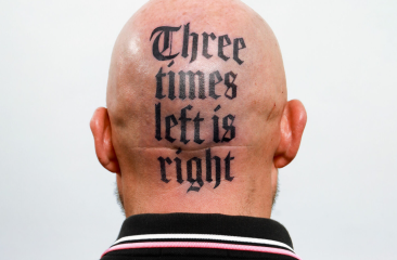 Kaalgeschoren achterhoofd van een persoon met een opvallende tatoeage in gotisch lettertype waarop staat: "Three times left is right".