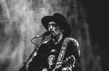 Zwart-witfoto van een man met hoed die zingt en gitaar speelt op een podium, met een expressieve blik en een shirt met een portret erop.