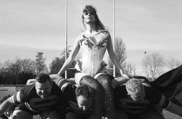 Zangeres in glitteroutfit zit op rugbyscrum van drie mannen met Superman-shirts op een sportveld