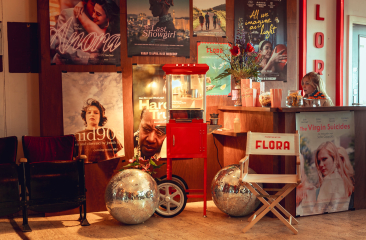 Een sfeervol ingerichte filmtheaterlobby met filmposters aan de muur, een retro popcornmachine, twee discoballen, een witte regisseursstoel met "FLORA" erop, en een medewerker achter de balie.