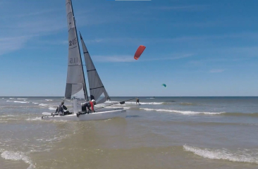 Catamaran met opgestoken zeilen vaart de zee op, met kitesurfers verderop in beeld onder blauwe lucht