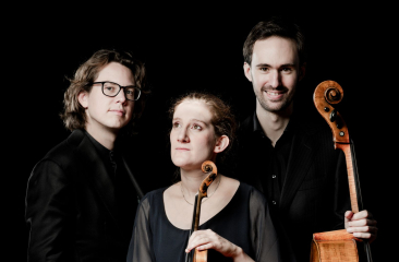 Drie musici poseren met strijkinstrumenten tegen zwarte achtergrond, twee met cello en één met viool in het midden
