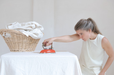 Vrouw in witte outfit strijkt geconcentreerd een tafelkleed met een klein oranje strijkijzer naast een rieten wasmand