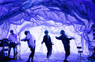 Vier mensen dansen onder een kunstzinnig verlicht decor dat lijkt op een grot van paarse doeken, met een muzikant links die live muziek maakt.
