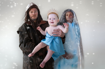 Man en vrouw in winterse kleding houden baby met blauwe doek en kroon vast in vallende sneeuw.