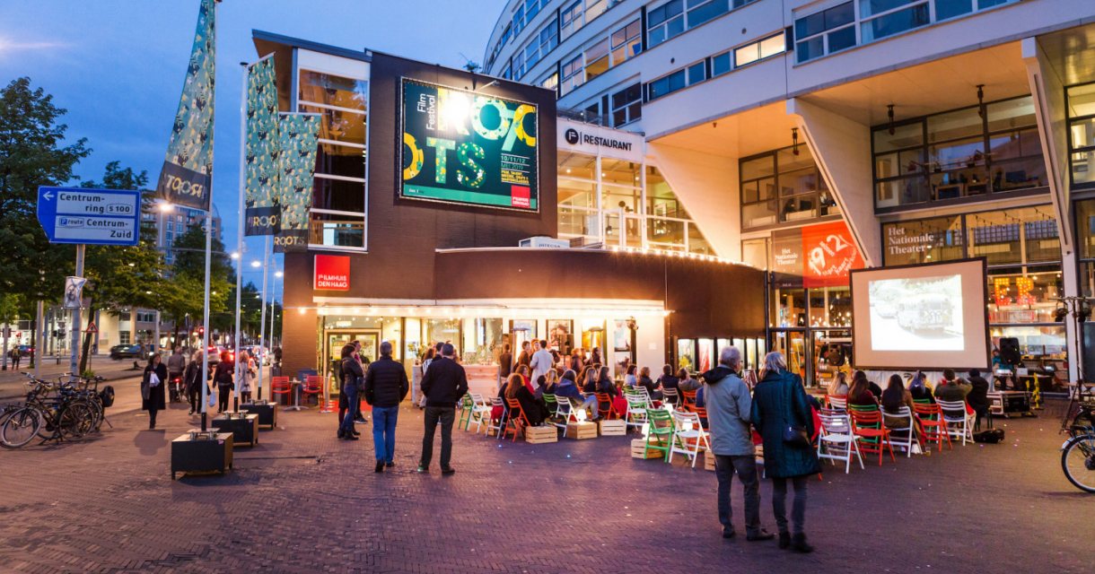 Mensen voor de sfeervol verlichte ingang van het Filmhuis tijdens het Roots Film Festival Mensen voor de sfeervol verlichte ingang van het Filmhuis tijdens het Roots Film Festival