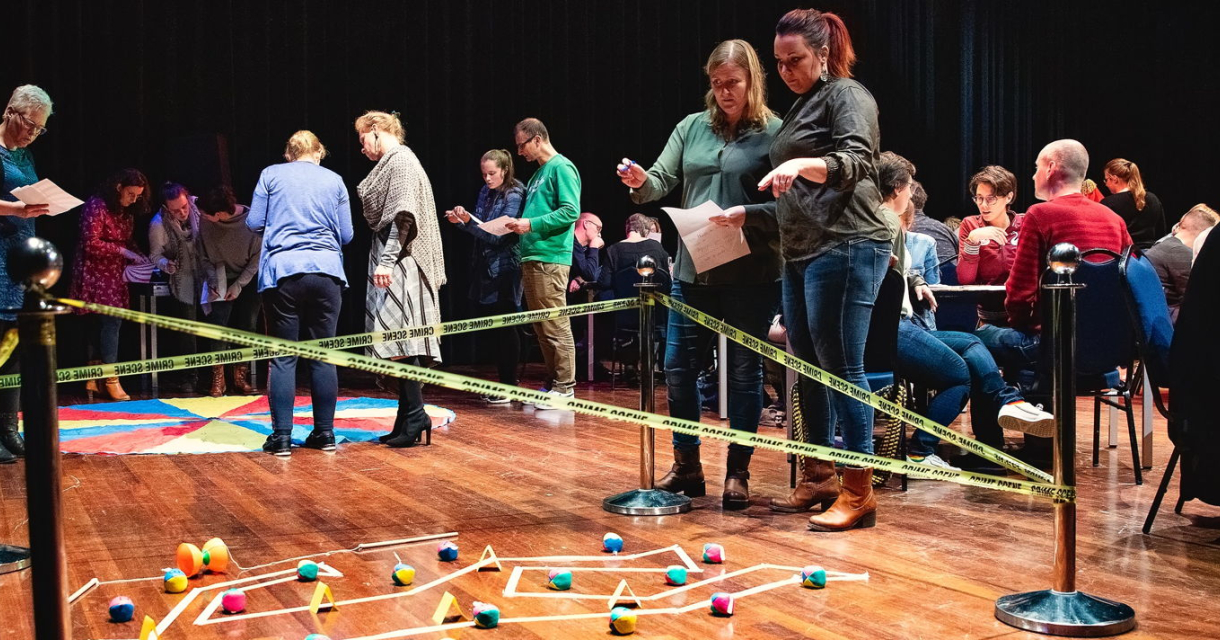 Foto van bezoekers van het Moordmysterie. Bezoekers kijken naar een crimescene op de vloer terwijl andere bezoekers aan tafels overleggen. Foto van bezoekers van het Moordmysterie. Bezoekers kijken naar een crimescene op de vloer terwijl andere bezoekers aan tafels overleggen.