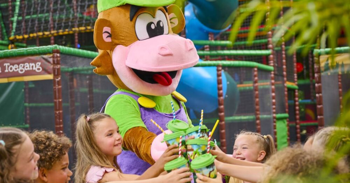 Een vrolijke mascotte in de vorm van een aap deelt bekers uit aan lachende kinderen in een kleurrijke indoor speeltuin. Een vrolijke mascotte in de vorm van een aap deelt bekers uit aan lachende kinderen in een kleurrijke indoor speeltuin.