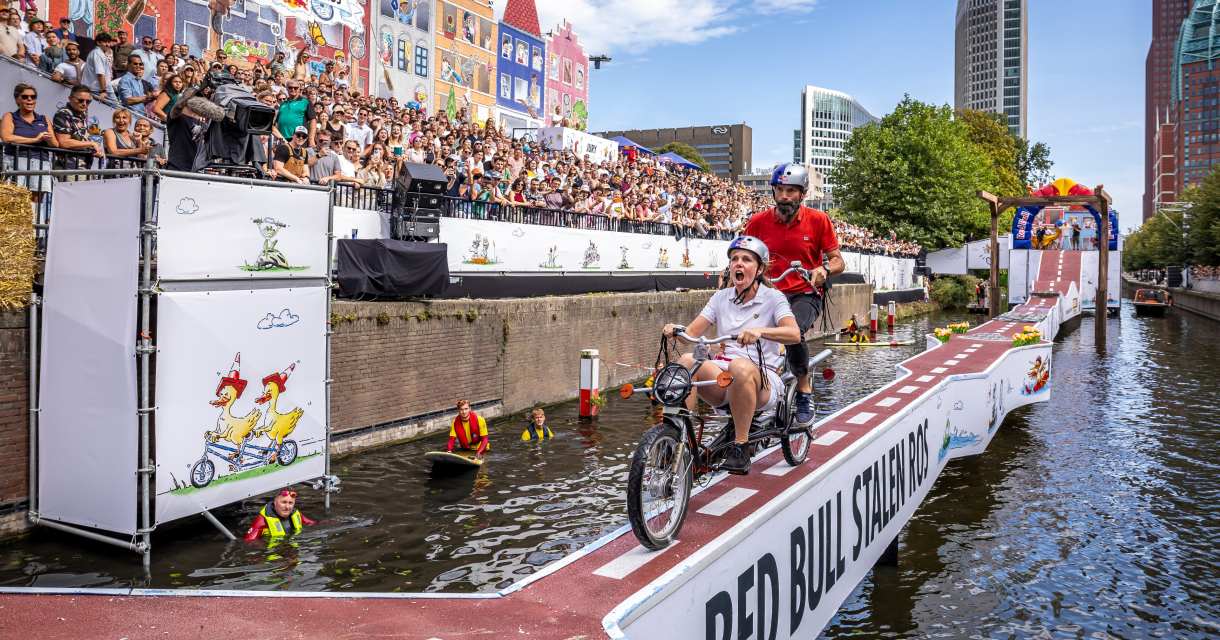 Twee deelnemers op een tandemfiets racen over een schans tijdens Red Bull Showrun in een drukbezocht, kleurrijk Den Haag. Twee deelnemers op een tandemfiets racen over een schans tijdens Red Bull Showrun in een drukbezocht, kleurrijk Den Haag.