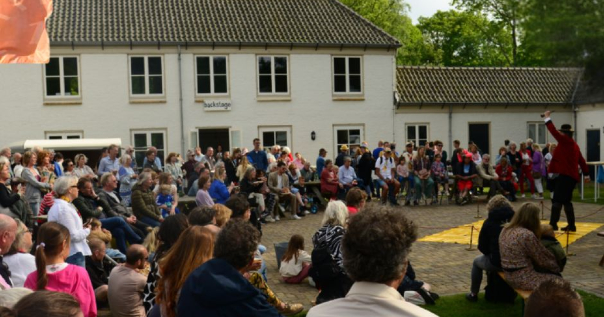 Buitenvoorstelling voor een groot publiek op een binnenplaats bij een wit gebouw, met een artiest die optreedt voor een aandachtig toekijkende menigte. Buitenvoorstelling voor een groot publiek op een binnenplaats bij een wit gebouw, met een artiest die optreedt voor een aandachtig toekijkende menigte.